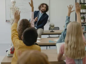 A classroom of kids raising their hands while the teacher holds a pointer.