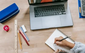 Student writing notes in a notebook with a laptop and school supplies on the desk.