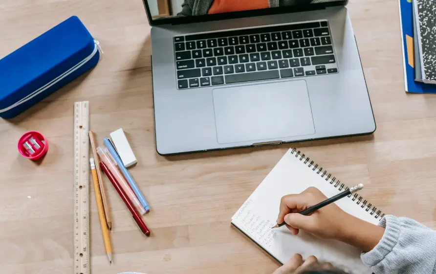 Student writing notes in a notebook with a laptop and school supplies on the desk.