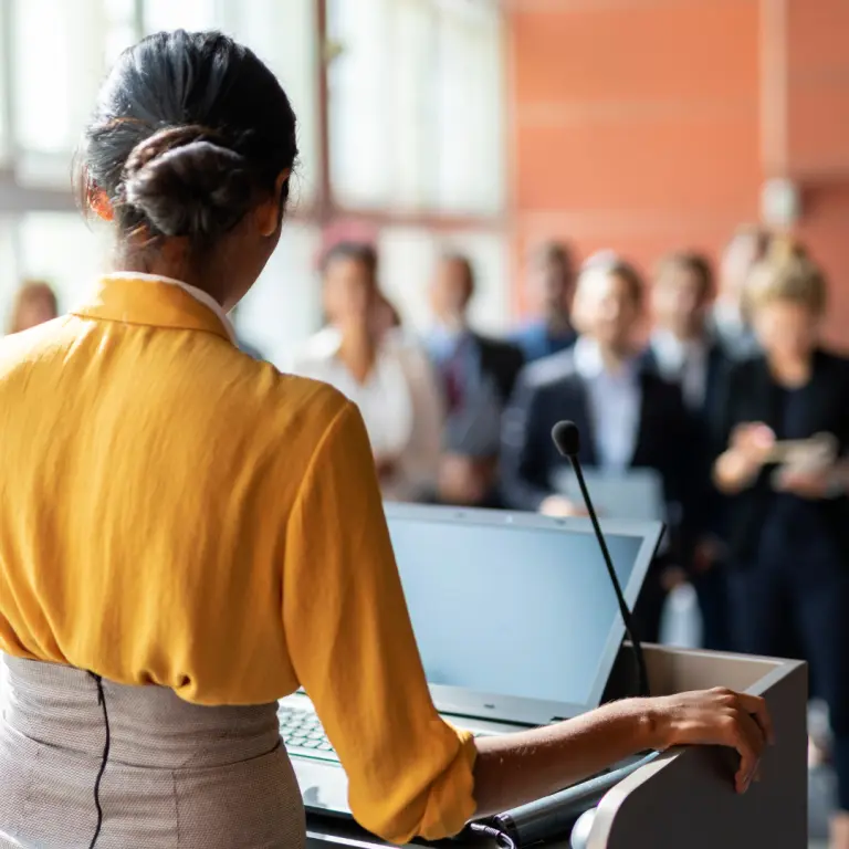 Woman in a yellow blouse speaking at a podium to an audience.